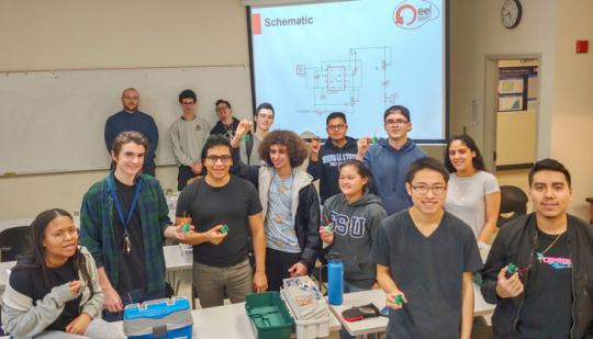 EE 110 students along with soldering mentors against the white board, from left to right: Joseph Haun, David Story and Adam Ortega